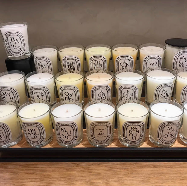 Display of Diptyque candles in clear glass jars with white labels, showcasing 24 visible scents including Baies, Tubereuse, Patchouli, and Gardenia, arranged in rows with varied wax colours