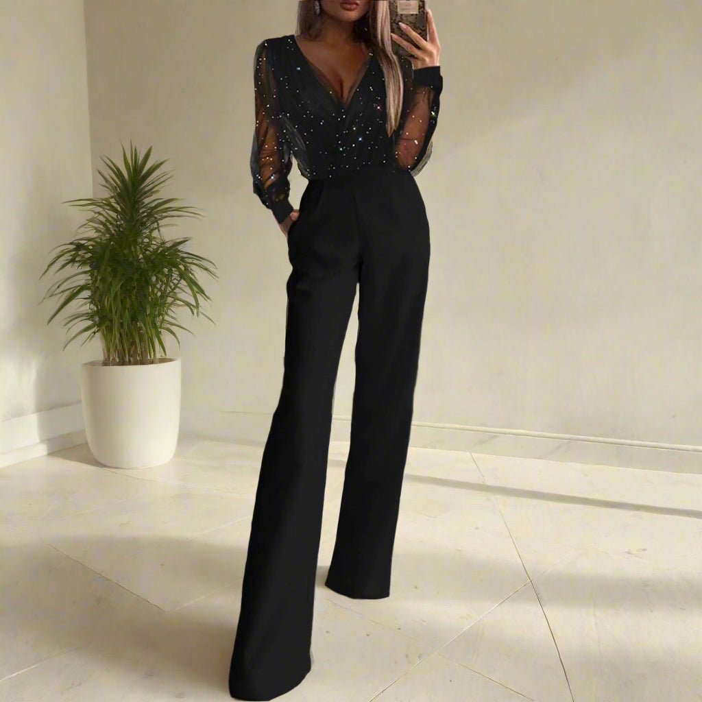 Stylish woman wearing a black jumpsuit with wide-legged pants and sheer, embellished long sleeves, taking a mirror selfie in a room with a potted plant and tiled floor, modern, chic, and versatile