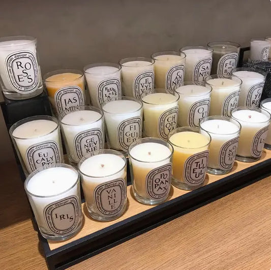 Display of Diptyque candles in clear glass jars with white labels, showcasing 24 visible scents including Baies, Tubereuse, Patchouli, and Gardenia, arranged in rows with varied wax colours