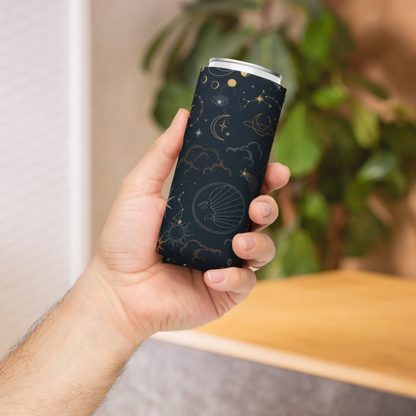 Hand holding a black can cooler with celestial-themed prints in gold and white—featuring stars, planets, clouds, and an eye, wrapped around a slim can—stylish, insulated, and available in two sizes.