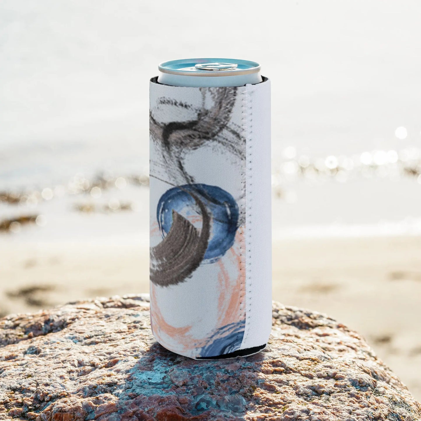 Can wrapped in an abstract-designed sleeve featuring swirls and strokes in blue, peach, and black, placed on a rock by the beach—modern, artistic, and perfect for casual drink accessories