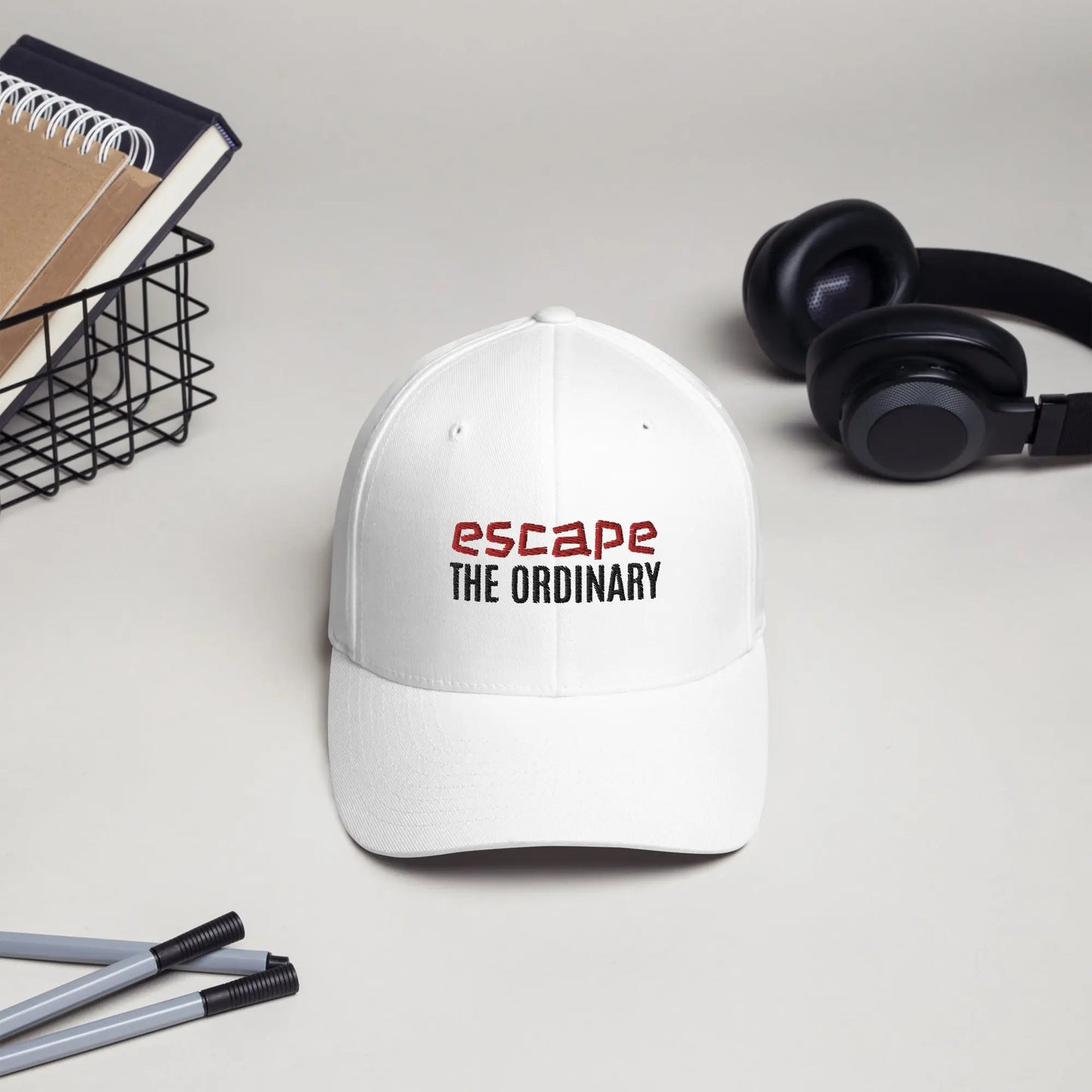 White baseball cap featuring embroidered text reading ‘escape THE ORDINARY’ in red and black, placed beside black headphones and stationery—modern, stylish, and perfect for casual wear.