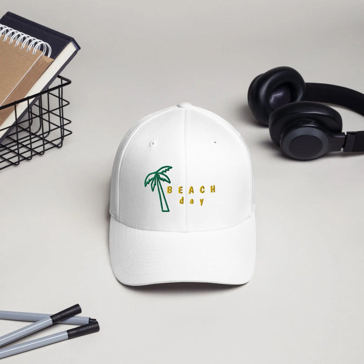 White twill cap featuring embroidered palm tree and ‘BEACH day’ text in yellow, placed beside a black wire basket, headphones, and stationery—casual, stylish, and perfect for summer outfits.