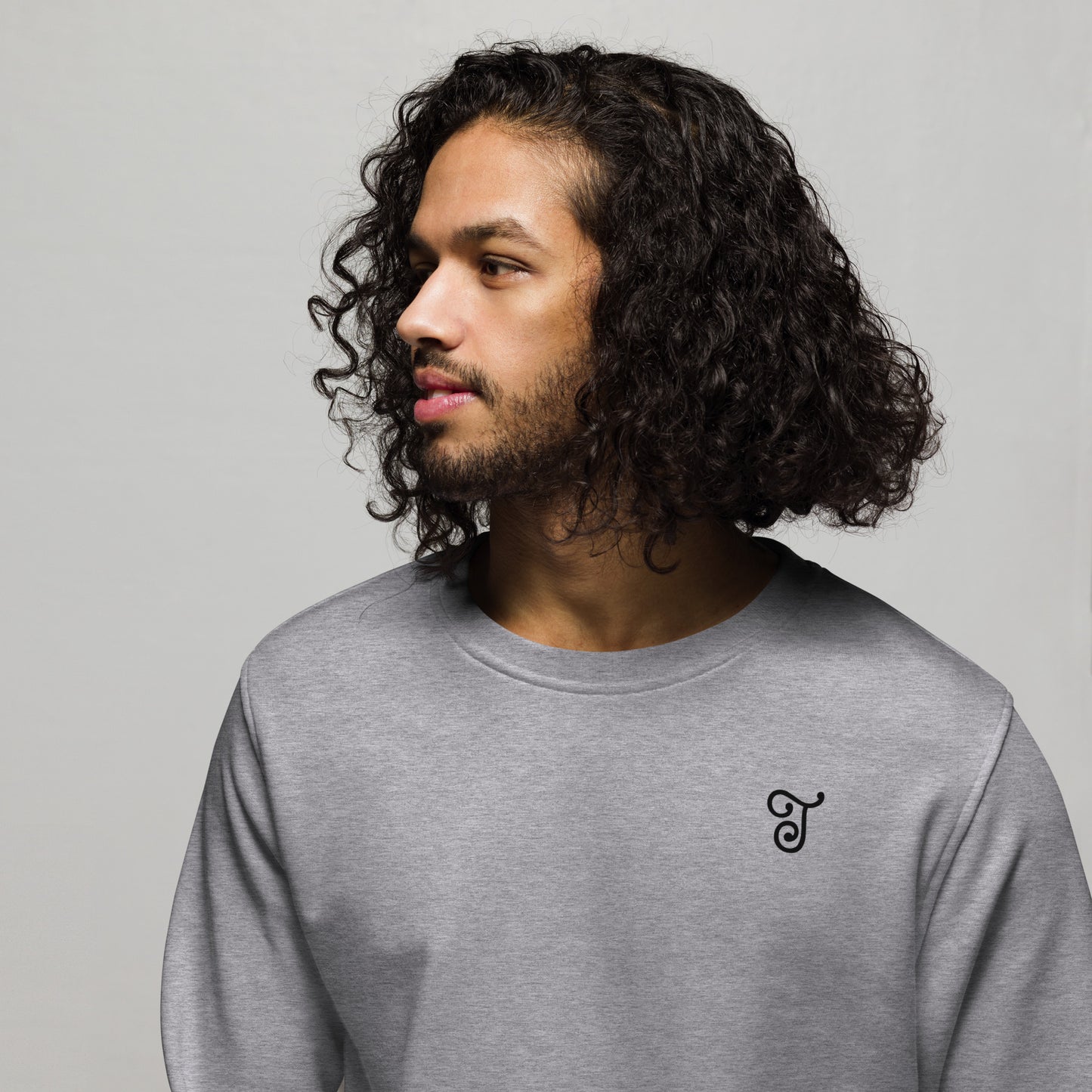 Grey sweatshirt featuring an embroidered letter ‘T’ on the left chest, worn with beige pants, seated on a black stool against a white background—modern, stylish, and available in multiple colors.