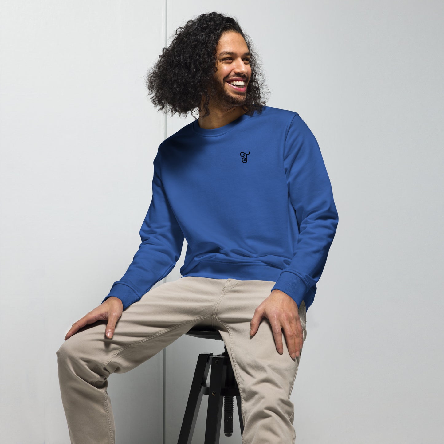Royal blue sweatshirt featuring an embroidered letter ‘T’ on the left chest, worn with beige pants, seated on a black stool against a white background—modern, stylish, and available in multiple colors.