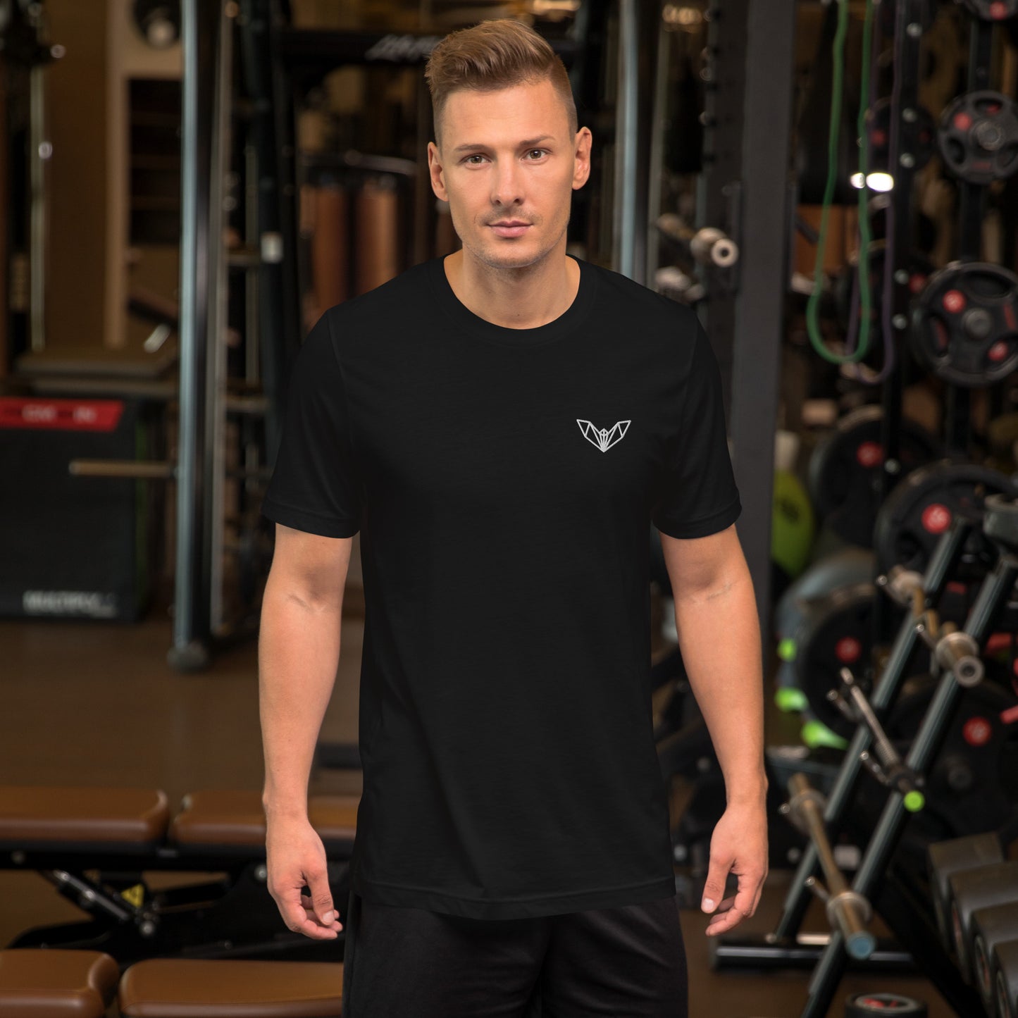 Black athletic t-shirt featuring a sharp geometric bat logo in white on the left side of the chest, worn in a gym environment with various exercise equipment—sleek, edgy, and perfect for fitness and casual wear.