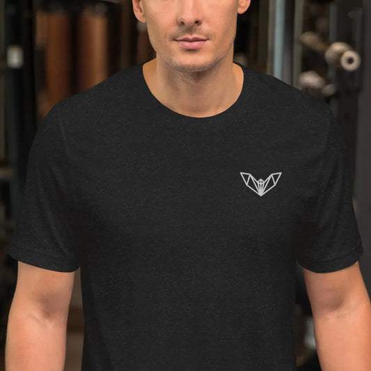 Black athletic t-shirt featuring a sharp geometric bat logo in white on the left side of the chest, worn in a gym environment with various exercise equipment—sleek, edgy, and perfect for fitness and casual wear.