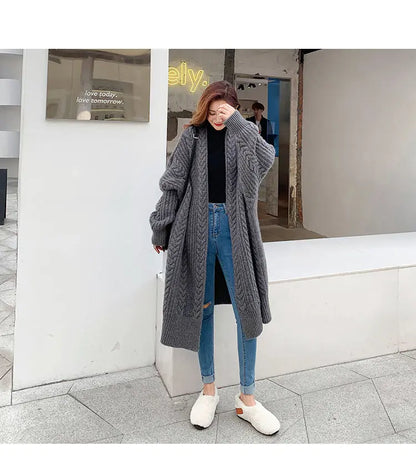 A woman stands outdoors wearing a long, oversized chunky-knit gray cardigan over a black top and blue jeans, with white slip-on shoes. The background shows a modern white wall, glass storefront window, and tiled pavement.