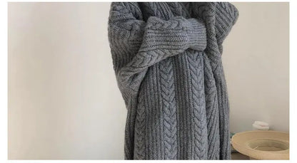 A close-up of a chunky gray cable-knit sweater draped on a mannequin or person, showing the thick knit texture and oversized sleeve detail against a plain indoor wall.