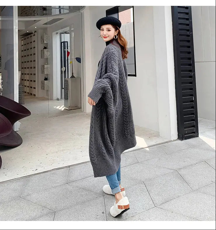 A woman wearing a long, chunky gray cable-knit cardigan, blue jeans, white wedge sandals, and a black beret stands on a tiled sidewalk in front of a modern storefront.