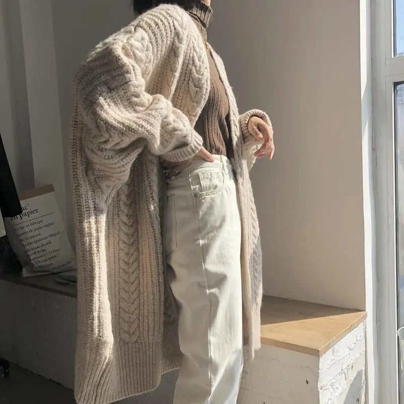 Person wearing a large beige knitted cardigan in a room with a window.
