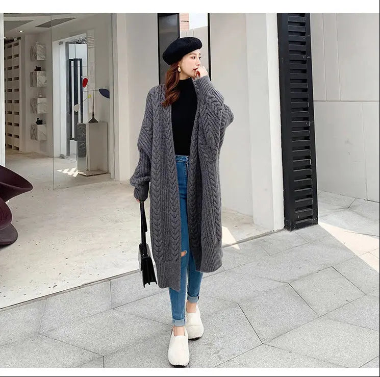 A woman stands outdoors wearing a long, oversized gray cable-knit cardigan over a black top and blue jeans, with white slip-on shoes and a black beret.