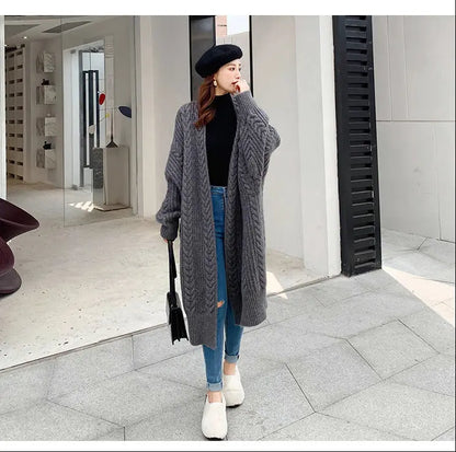 A woman stands outdoors wearing a long, oversized gray cable-knit cardigan over a black top and blue jeans, with white slip-on shoes and a black beret.