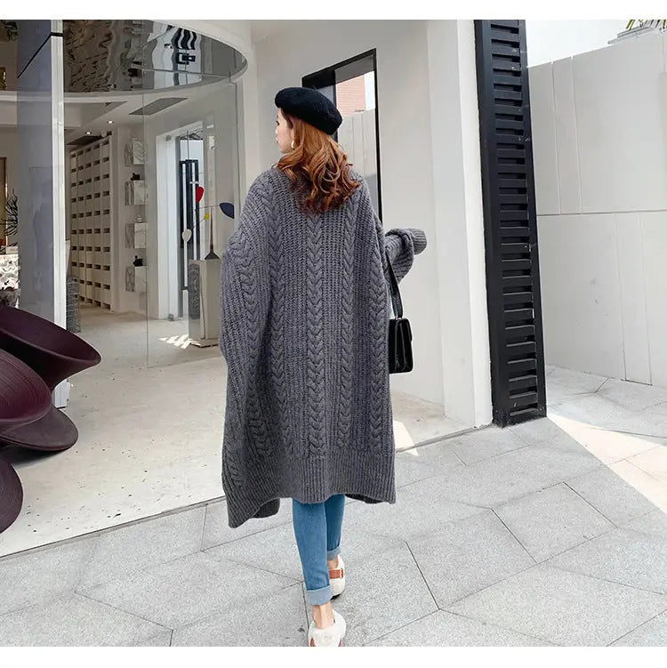 A woman wearing a long, chunky cable-knit gray cardigan, blue jeans, and a black beret walks past a modern glass storefront.