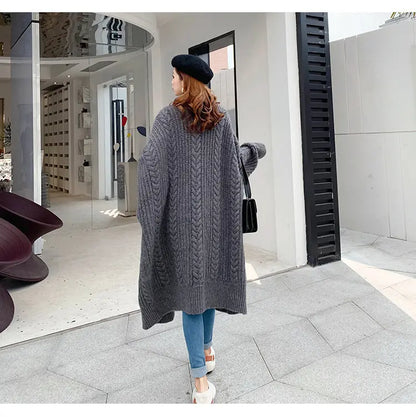 A woman wearing a long, chunky cable-knit gray cardigan, blue jeans, and a black beret walks past a modern glass storefront.