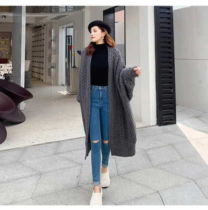 A woman stands outdoors wearing a long, chunky cable-knit gray cardigan over a black top and light blue ripped jeans, with white slip-on shoes and a black beret.