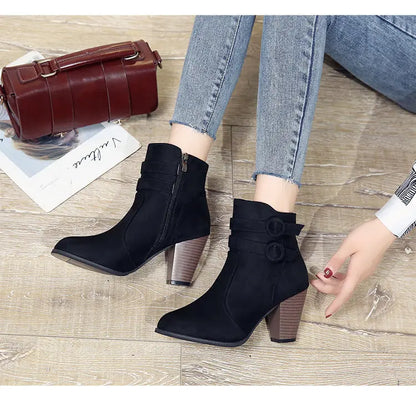 A pair of women's black ankle boots with chunky brown block heels and decorative side buttons, shown being worn with blue jeans on a light wood floor. A dark red handbag and magazine are visible in the background.