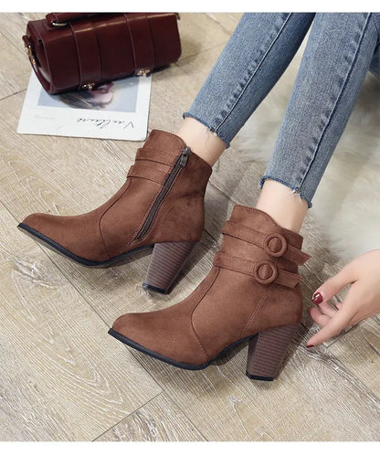 A pair of women's brown suede-look ankle boots with block heels, shown on a model wearing blue jeans. The boots feature a side zipper and double strap details with round buckle embellishments.