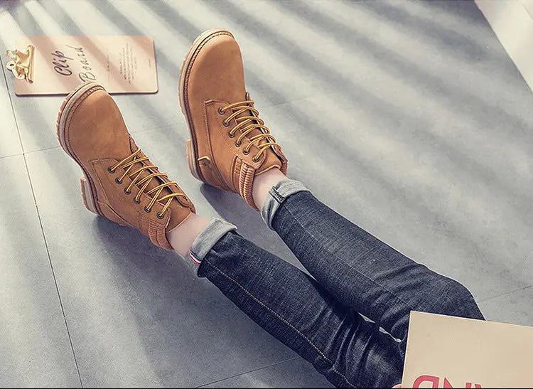 A pair of tan lace-up ankle boots worn with dark blue jeans, shown in two views: one standing on a gray floor and one lying on the floor with a book nearby.