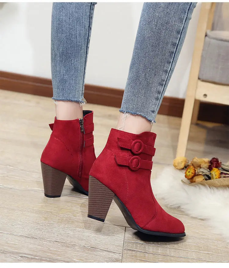 A person wearing red ankle boots with double buckle straps and brown block heels, paired with light blue frayed-hem jeans, standing on a light wood floor.