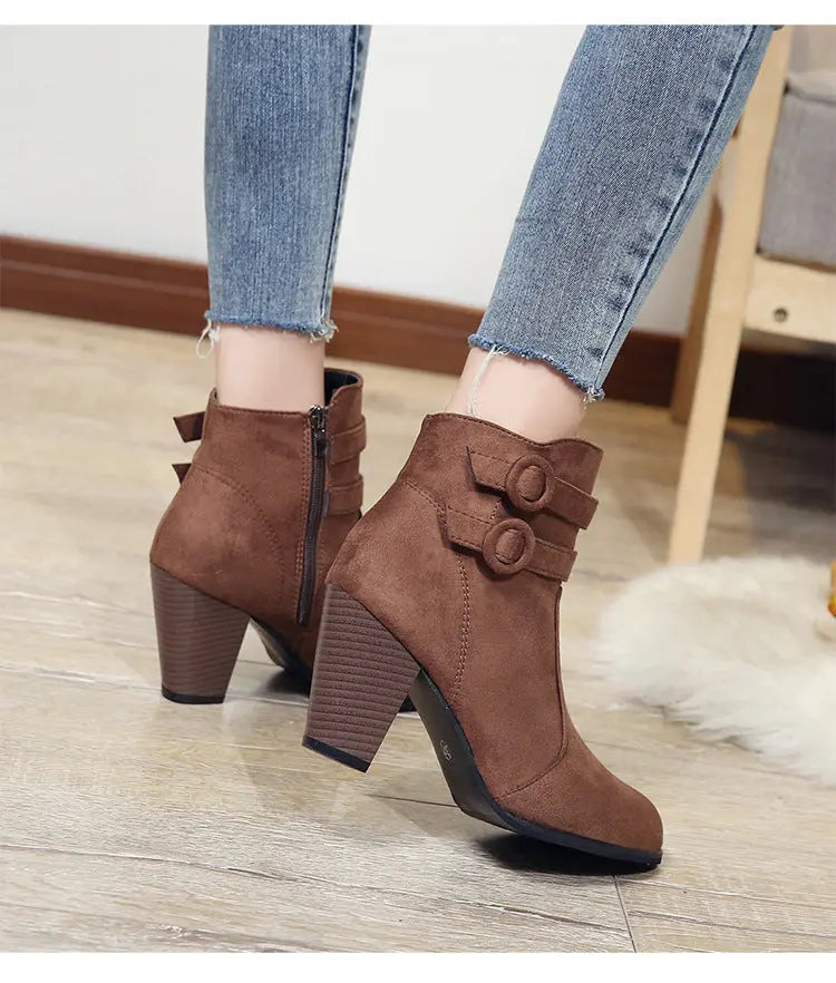 A pair of women's brown suede-look ankle boots with block heels, shown being worn with frayed-hem blue jeans on a light wood floor.