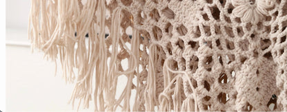 Close-up of a cream-colored crocheted or knitted textile with long fringe and openwork floral-like patterns.