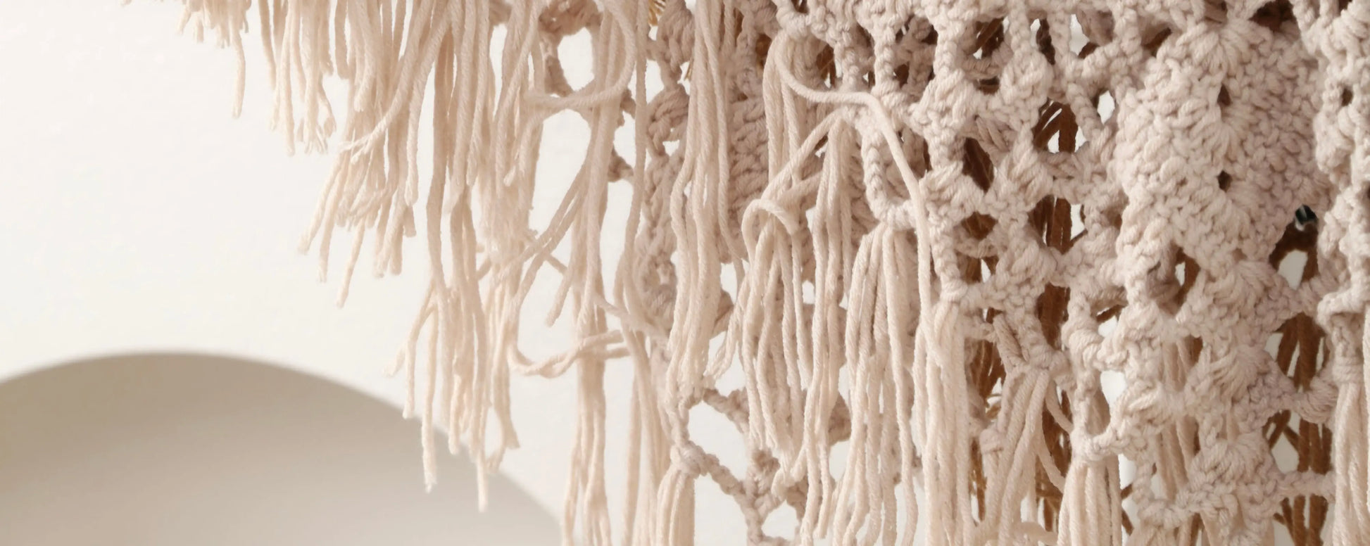 Close-up of a cream macrame or knotted textile with long fringe and openwork crochet-like patterns against a light neutral background.