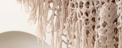 Close-up of a cream macrame or knotted textile with long fringe and openwork crochet-like patterns against a light neutral background.