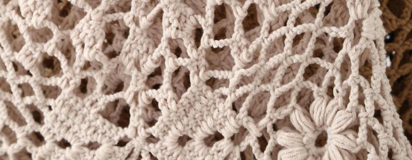 Close-up of a beige crochet or lace textile with an openwork floral pattern, showing the texture and stitch detail.