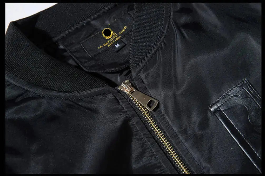 Close-up of a black nylon bomber jacket showing the ribbed collar, a gold-tone zipper, and a black neck label with a gold logo.