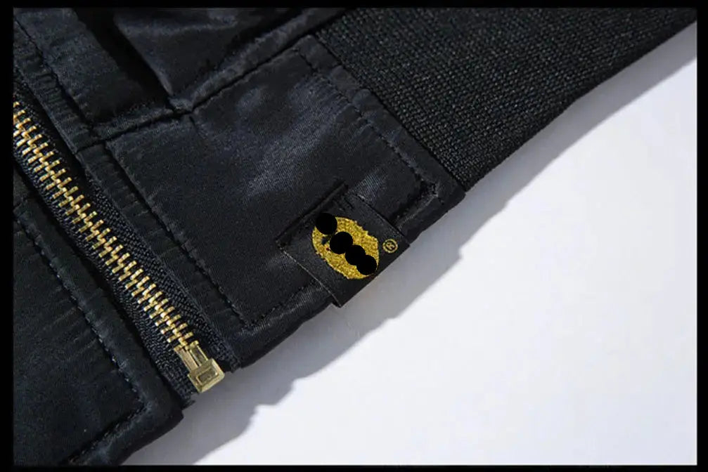Close-up of a black jacket with a gold zipper and a small gold patch featuring a stylized face logo, set against a white background.