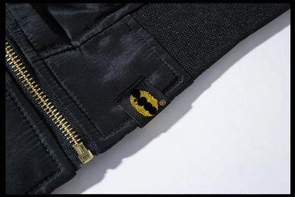 Close-up of a black jacket with a gold zipper and a small gold patch featuring a stylized face logo, set against a white background.
