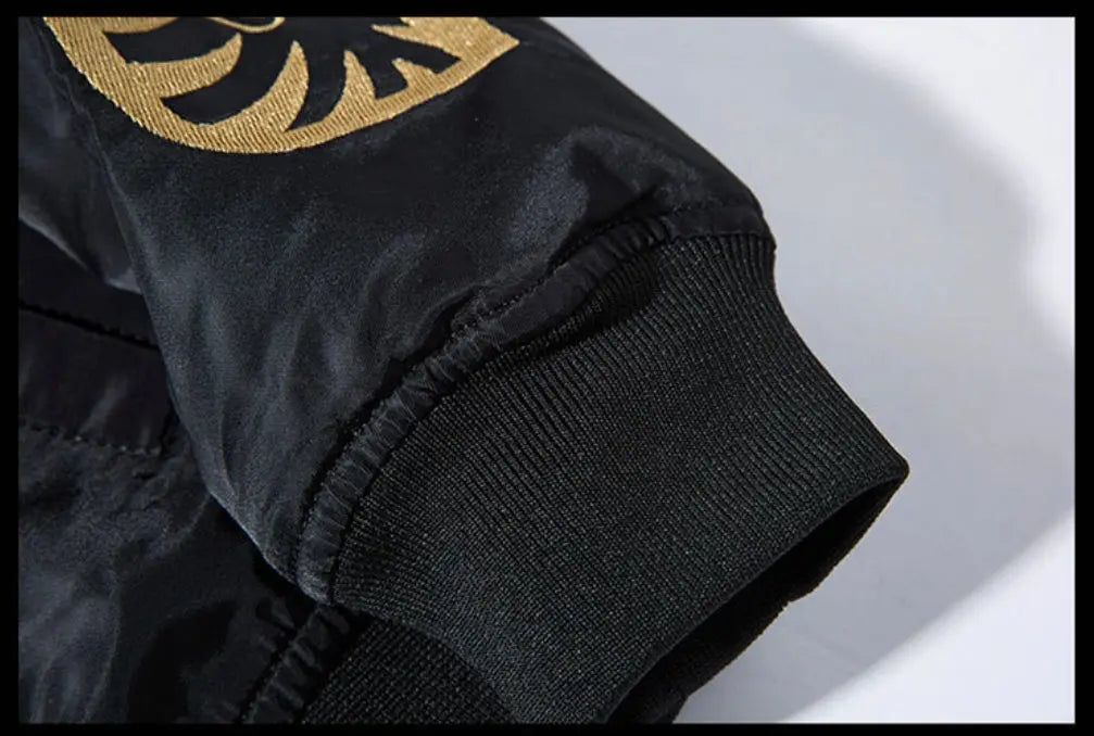 Close-up detail of a black bomber jacket sleeve featuring a ribbed knit cuff and a gold embroidered circular emblem on the upper arm.