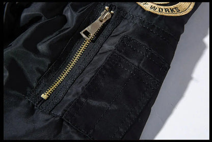 Close-up of a black jacket or pants fabric with a gold zipper, a gold-and-black patch reading WORKS, and visible stitching on a white background.