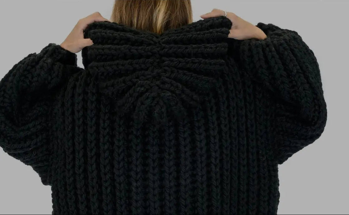 A person viewed from behind wearing a chunky black knit hooded sweater, holding the hood up with both hands against a plain gray background.