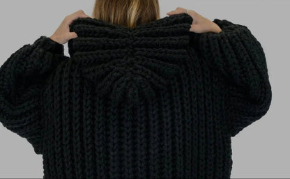 A person viewed from behind wearing a chunky black knit hooded sweater, holding the hood up with both hands against a plain gray background.