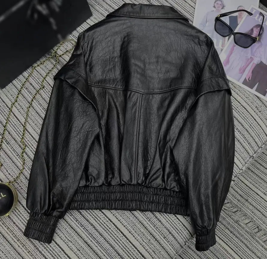 Black leather jacket shown from the back, laid flat on a gray striped textured surface with sunglasses and a magazine page nearby.