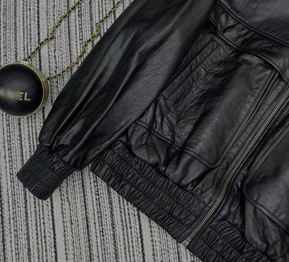 A close-up flat lay of a black leather jacket with elasticized cuffs and a zippered pocket, placed on a gray textured surface next to a round black pendant on a chain.