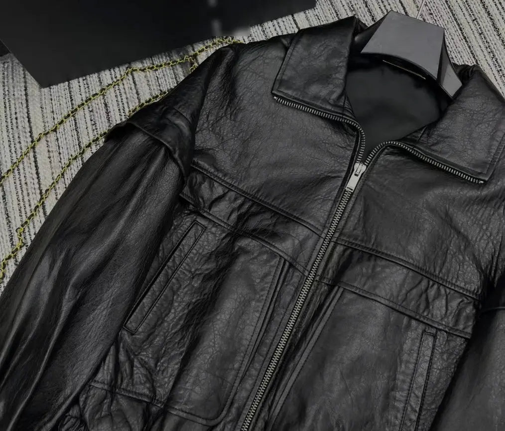 A close-up of a black leather jacket with a front zipper and chest pockets, laid flat on a textured gray and white patterned surface.