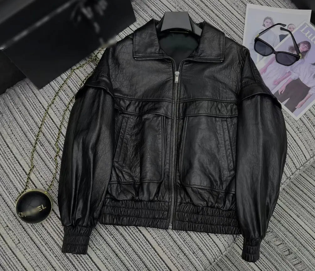 A black leather jacket with a high collar, front zipper, and elasticized cuffs and hem, laid flat on a gray striped textured surface with sunglasses and a magazine nearby.