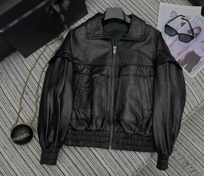 A black leather jacket with a high collar, front zipper, and elasticized cuffs and hem, laid flat on a gray striped textured surface with sunglasses and a magazine nearby.