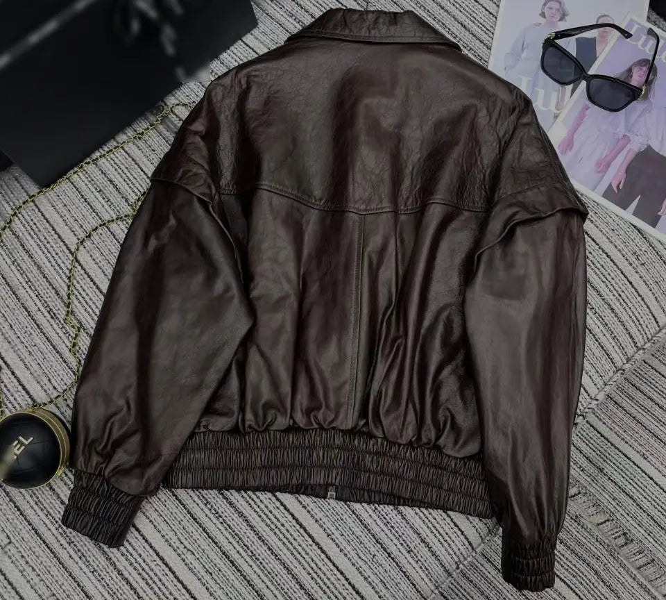 A dark brown faux leather jacket shown from the back, laid flat on a gray and white striped carpet with fashion accessories like sunglasses and a watch nearby.