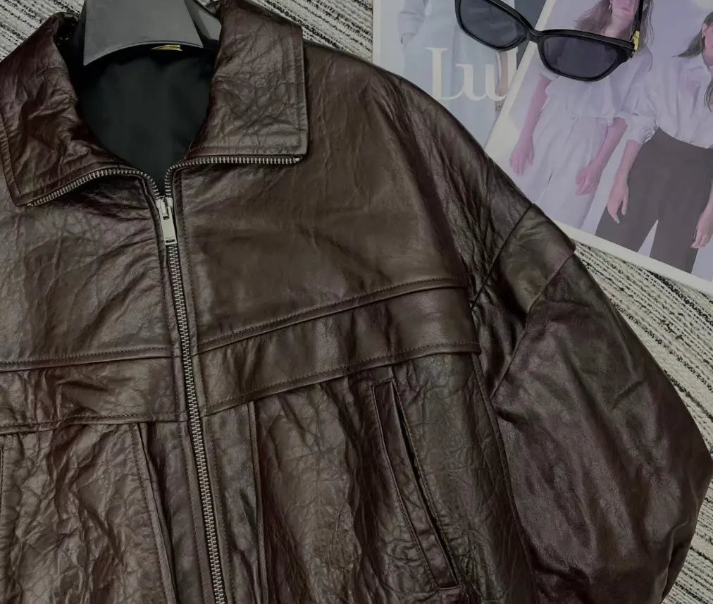 A dark brown crinkled faux leather jacket with a front zipper, laid flat on a textured gray and white striped blanket with a magazine and sunglasses in the background.