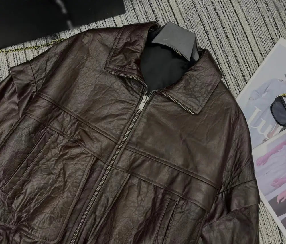 A dark brown crinkled leather jacket with a front zipper and stand collar, laid flat on a textured gray and white striped rug with a magazine partially visible on the right.