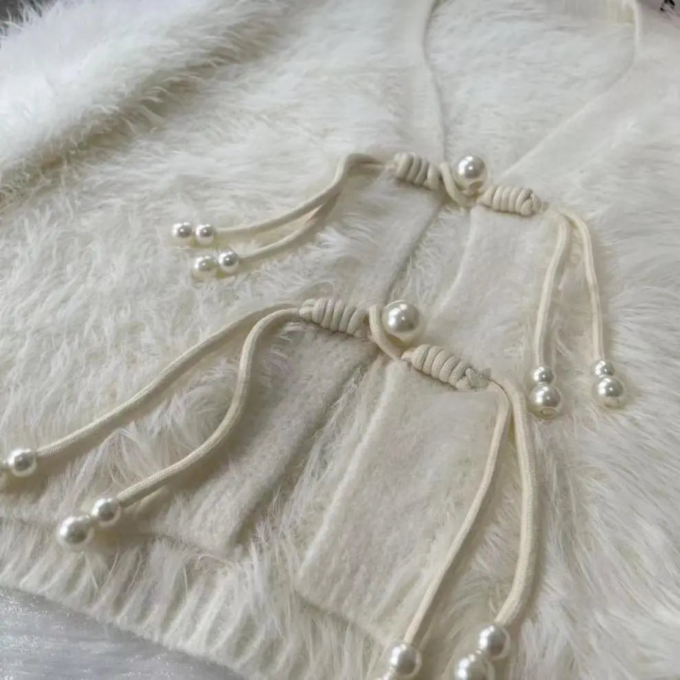 Close-up of a cream-colored knit garment featuring decorative cord ties with faux pearl beads, laid flat on a white faux fur surface.