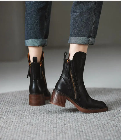 Black leather boots with brown soles worn with rolled-up jeans on a neutral background