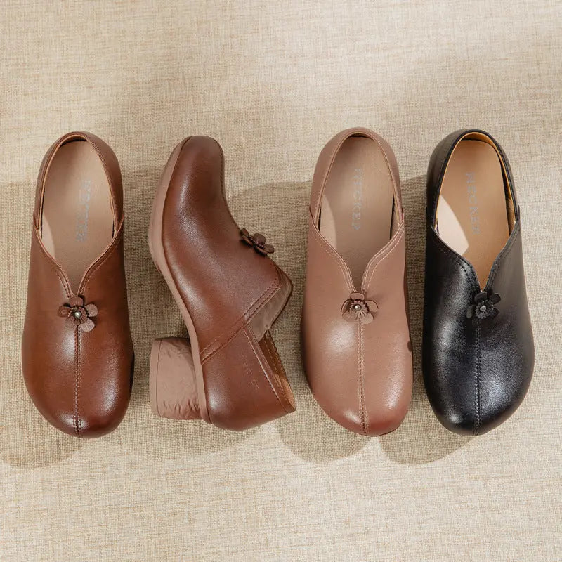 Four women's slip-on mule shoes arranged on a beige textured fabric background. The set includes brown, medium brown, taupe, and black versions, each with a small decorative flower detail at the front and a low block heel.