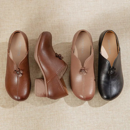 Four women's slip-on mule shoes arranged on a beige textured fabric background. The set includes brown, medium brown, taupe, and black versions, each with a small decorative flower detail at the front and a low block heel.