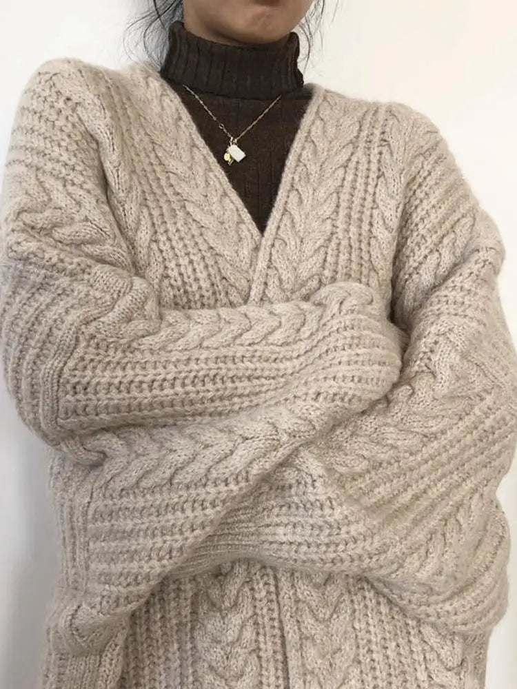 A close-up of a person wearing an oversized chunky cable-knit cardigan in a warm beige/taupe color over a dark brown turtleneck sweater, with a delicate gold necklace visible.