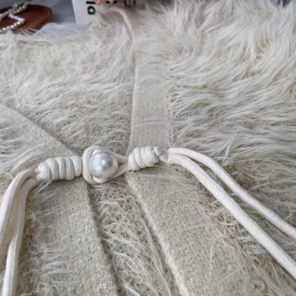 Close-up of a cream-colored knit garment with a knotted cord detail and a central faux pearl, laid flat on a fluffy cream faux-fur surface.